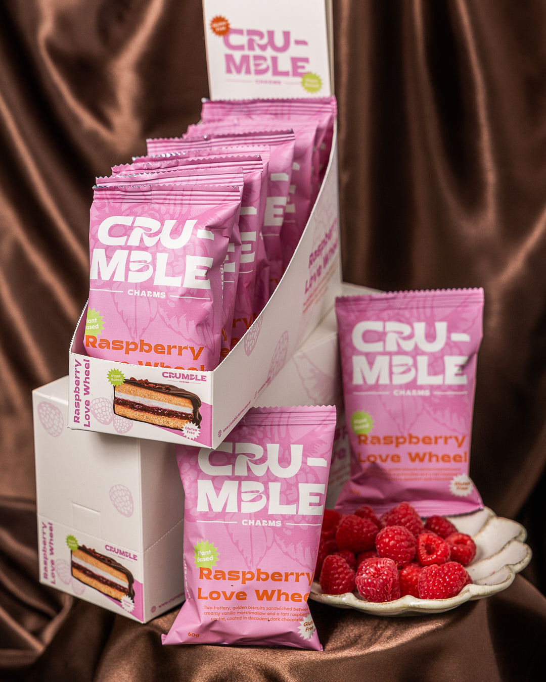 Display of Crumble Charms Raspberry Love Wheel snack packs with fresh raspberries on a chocolate satin background