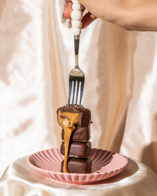 Fork lifting a gooey caramel chocolate bar over stacked Crumble Charms treats on a pink plate